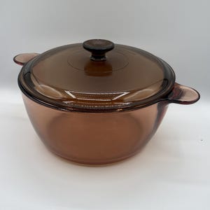 May include: A round, amber-colored glass casserole dish with a matching lid. The dish features two integrated side handles and a central knob on the lid. The translucent brown glass has a smooth, polished finish throughout the entire piece.