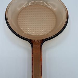Könnte beinhalten: Eine runde Pfanne aus bernsteinfarbenem Glas mit einem langen Griff. Die Bratfläche weist ein dezentes, erhabenes Gittermuster auf. Der Griff ist mit dem Text "VISIONS" und "CORNING" sowie "USA" und weiteren kleinen Markierungen geprägt.