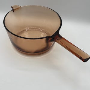 Pode incluir: Uma panela de vidro translúcido na cor âmbar com corpo redondo, uma alça longa e um pequeno bico vertedor na borda. O vidro tem um acabamento liso e polido com um tom castanho quente e consistente em todo o recipiente.