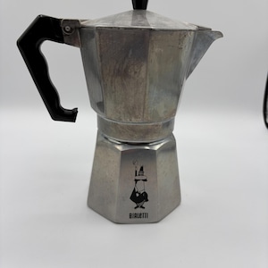 May include: A silver-coloured aluminium stovetop espresso maker with an octagonal base and a black plastic handle and lid knob. The lower chamber features a black graphic logo of a person with a raised arm and the text BIALETTI printed below it.