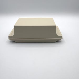 Può includere: Un burriera rettangolare di colore beige con coperchio abbinato. La base presenta un bordo leggermente più largo e il coperchio ha una superficie superiore liscia e piatta. Il design è semplice e funzionale, realizzato in materiale solido e opaco.