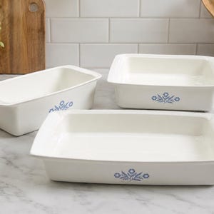 Puede incluir: Tres fuentes de horno de cerámica blanca de diversas formas y tamaños, incluyendo un molde rectangular para pan y dos fuentes rectangulares. Cada pieza presenta un pequeño motivo floral azul centrado en las paredes laterales exteriores.