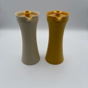 May include: Two vintage, hourglass-shaped kitchen dispensers. One is a muted beige, the other a mustard yellow. Both have a matching yellow lid with a small spout and a twist-top closure. The dispensers are made of plastic.