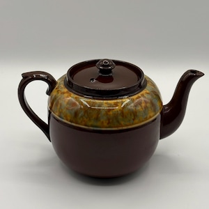 May include: A brown ceramic teapot with a lid and handle. The teapot has a rounded base and a textured, multi-coloured band around the middle. The lid is dark brown with a small knob. The spout and handle are also dark brown.