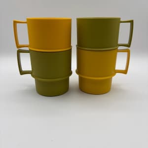 Puede incluir: Cuatro tazas apiladas en tonos amarillo y verde oliva. Cada taza tiene un asa rectangular y una base acanalada. Las tazas están dispuestas en dos pilas de dos, con los colores alternándose en cada pila.