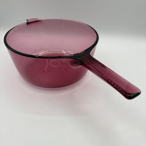 Könnte beinhalten: Ein durchscheinender magentafarbener Glasstieltopf mit einer runden Schale und einem einzelnen, länglichen Griff, der seitlich herausragt. Das Glas hat eine glatte, polierte Oberfläche mit einem tiefen violett-rosa Farbton und einem klaren Boden.