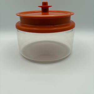 Peut inclure: Un récipient de stockage cylindrique transparent doté d'un couvercle en plastique orange vif. Le couvercle présente une poignée centrale en forme de bouton et un rebord large et plat qui s'ajuste sur la base transparente, formant un récipient.