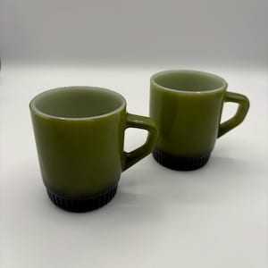 May include: Two olive green mugs with black ribbed bases and handles. The mugs have a cylindrical shape and a white rim. The mugs are made of a glossy material and are set against a white background. The mugs are likely vintage or retro.