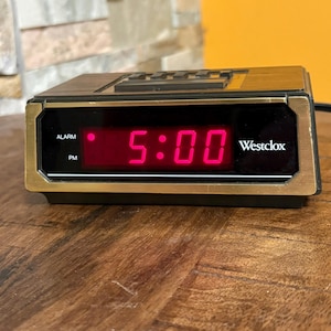 May include: A retro Westclox digital alarm clock showing 5:00 PM in red digits. The clock has a black body with gold-coloured trim and buttons on top. The word "Westclox" is printed on the right side of the display.