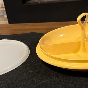Puede incluir: Un plato para servir amarillo dividido en tres secciones, con un asa central y una tapa ovalada blanca. El plato está diseñado para servir alimentos y la tapa está separada.