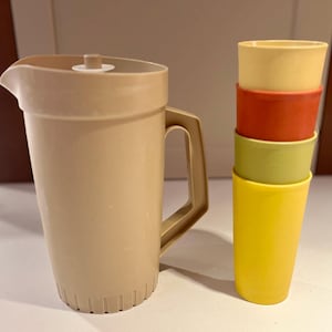 Puede incluir: Una jarra de plástico beige con tapa y asa está junto a una pila de cuatro vasos de plástico de colores. Las tazas son amarillas, verdes, rojas y amarillas. El conjunto es probablemente para servir bebidas.