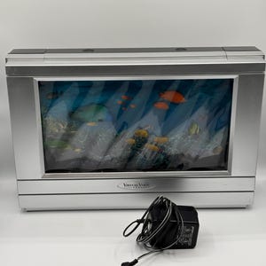 May include: A rectangular silver Virtual Vision electronic aquarium display featuring a printed underwater scene with orange and yellow fish, coral, and rocks. The device includes a black power adapter with a cord positioned in the foreground.