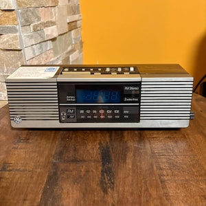 May include: A retro silver and brown FM stereo clock radio with a digital display showing the time. The radio has a silver front with horizontal lines, a black control panel, and a wood-grain top. The radio has FM and AM radio settings.