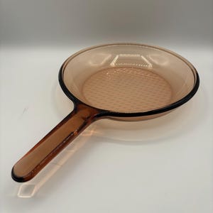 Può includere: Una padella di vetro trasparente color ambra con un lungo manico. La padella ha una superficie di cottura strutturata e un bordo nero. Il manico è un'estensione del corpo della padella. La padella è progettata per cucinare.