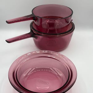 Puede incluir: Juego de tres piezas de utensilios de cocina Vision vintage en color rubí intenso. El juego incluye dos cacerolas con asas y una fuente redonda para horno. El logotipo de Vision es visible en la fuente.
