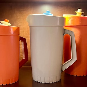 May include: Three vintage plastic jugs in orange and beige, with matching lids and handles. The jugs are cylindrical with a ridged base. The lids have small, colourful accents. The jugs are displayed on a wooden shelf.