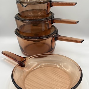 Puede incluir: Un juego de utensilios de cocina de vidrio color ámbar, que incluye tres cacerolas de diferentes tamaños apiladas y una sartén. Las cacerolas tienen mangos largos y tapas. La sartén tiene una superficie de cocción texturizada.