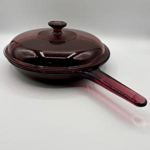Puede incluir: Una sartén de vidrio rojo rubí vintage con tapa a juego. La sartén tiene un mango largo y una superficie de cocción redonda. La tapa presenta un pomo y un patrón decorativo. Esta batería de cocina es ideal para cocinar y servir.