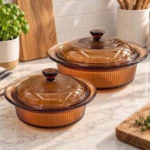 May include: Two round amber-coloured glass casserole dishes with matching lids. Each dish features a vertical ribbed pattern on the sides and a circular textured design on the centre of the lid, topped with a small, dark brown glass knob handle.