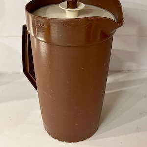 May include: A brown plastic pitcher with a handle and a lid. The pitcher has a wide opening and a spout for pouring. The lid is white with a brown knob. The pitcher is likely used for serving beverages.