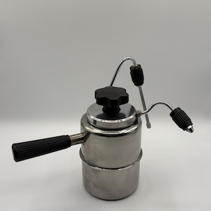 Puede incluir: Una cafetera espresso de acero inoxidable para estufa con cuerpo cilíndrico, mango lateral de plástico negro y una perilla negra en forma de estrella en la tapa. Dos varillas de vapor metálicas curvas con agarres negros sobresalen arriba.