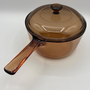 May include: A round, amber-coloured glass saucepan with a matching transparent glass lid. The cookware features a long, integrated glass handle extending from the side and a small, circular knob on the centre of the lid for easy removal.