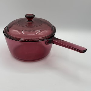 May include: A deep magenta-coloured glass saucepan with a matching lid. The lid features a central knob and a radial ribbed pattern. The saucepan has a single long handle extending from the side, and the entire piece is made of translucent glass.