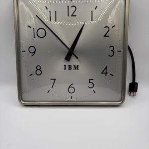 Può includere: Orologio quadrato con quadrante argentato e lancette e numeri neri. Il quadrante dell'orologio ha il logo IBM e un cavo di alimentazione nero. L'orologio è in una cornice di metallo argentato.
