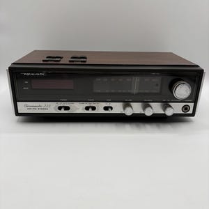 May include: A Realistic Chronomatic-223 AM-FM stereo receiver with a wood-grain top and a black front panel. The unit features a digital clock display, analog radio tuning dial, silver control knobs, and a headphone jack on the right side.