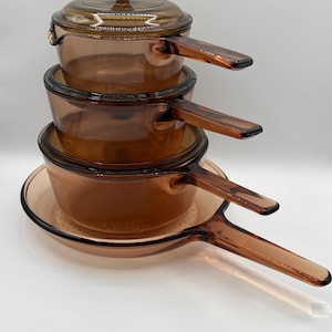Pode incluir: Um conjunto de quatro peças de cozinha em vidro cor de âmbar, incluindo três caçarolas com pegas integradas e uma tampa a condizer. O conjunto apresenta um tom castanho translúcido, bordos arredondados e um design consistente em todas as peças.