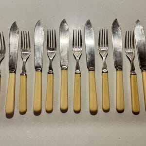 May include: A set of vintage silverware, including forks and knives, arranged on a light-colored surface. The knives have silver blades and cream-colored handles. The forks have silver tines and cream-colored handles. The silverware is likely for dining.