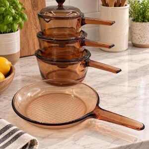 Peut inclure: Un ensemble d'ustensiles de cuisine en verre ambré comprenant trois casseroles emboîtables avec poignées, un couvercle en verre assorti et une poêle peu profonde avec un long manche. L'ensemble présente une finition lisse et translucide.