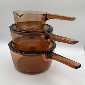 May include: Three amber glass saucepans stacked. Each pan has a handle and a lid. The smallest saucepan at the top has measurement markings. The pans are in a warm, brown hue.