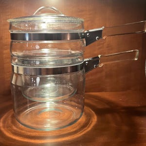 Puede incluir: Una olla de doble caldera de vidrio transparente con tapa y dos asas transparentes. El vidrio se mantiene unido con bandas de metal plateado. La olla de doble caldera está sobre una superficie de madera.