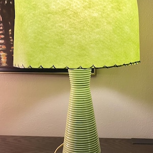 Może przedstawiać: Lampa stołowa w stylu retro z limonkowym abażurem i jasnozieloną, żebrowaną podstawą. Krawędź abażuru ma falisty brzeg z czarnymi przeszyciami. Podstawa stoi na czarnym metalowym trójnogu. Lampa jest podłączona.