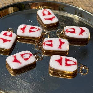 Puede incluir: Colgantes rectangulares blancos y rojos con forma de bikini, con bordes dorados y un pequeño lazo dorado para colgar. Los colgantes están dispuestos sobre una bandeja plateada.