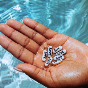 May include: A hand holding several small, white, pill-shaped charms with the word "love" written in black script. The charms have gold-colored accents and are set against a blue water background.