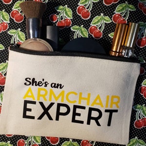 May include: A white canvas zippered pouch with a black zipper and black trim. The pouch has the text "She's an ARMCHAIR EXPERT" printed on it in black and yellow letters.