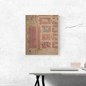 May include: Framed vintage map print with a detailed layout of a city, featuring pink and green building outlines on a beige background. The map is encased in a gold-colored frame, suitable for wall decor.