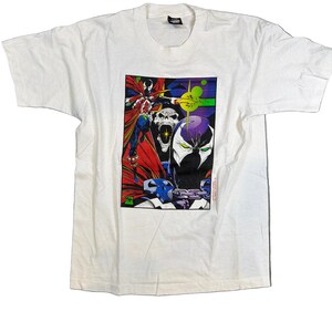 Puede incluir: Camiseta blanca con un gráfico colorido de personajes de cómic. El diseño presenta personajes con capa roja, una calavera y una figura con máscara morada. La camiseta tiene cuello redondo y mangas cortas.