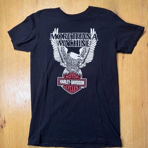 May include: Black t-shirt with a white eagle graphic and the text "MORE THAN A MACHINE" above and "MOTOR HARLEY-DAVIDSON CYCLES" below in a red and white emblem. The shirt is made of a soft material.