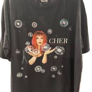 Pode incluir: Camiseta preta com um gráfico de Cher com cabelo ruivo, rodeada por bolhas. O texto "CHER believe" está em branco. A camiseta está pendurada em um cabide cinza.