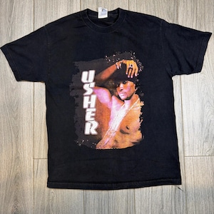 May include: Black t-shirt featuring a graphic of Usher. The image shows Usher wearing a hat and jewelry. The word "USHER" is printed vertically in white letters on the left side of the graphic. The shirt is made of a soft material.