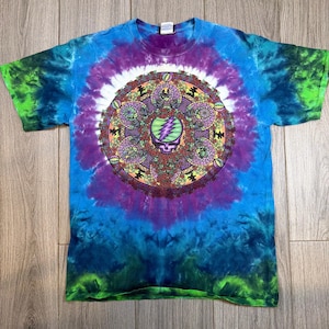 May include: A tie-dye t-shirt with a vibrant spiral design in shades of blue, purple, and green. The center features a detailed graphic with a skull and lightning bolt motif. The shirt is made of a soft material.