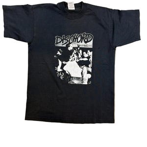 Puede incluir: Camiseta negra con el nombre de la banda "Dischord" en blanco, sobre un gráfico en blanco y negro de una multitud. La camiseta es de cuello redondo y manga corta.