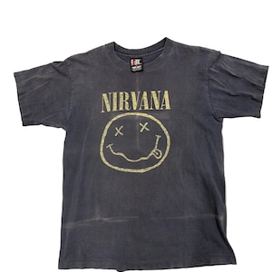 Peut inclure: T-shirt gris foncé avec le nom du groupe "NIRVANA" en lettres dorées au-dessus d'un smiley jaune avec des yeux barrés et une langue tirée. Le t-shirt a des manches courtes et un col rond.