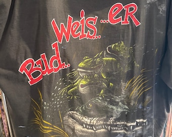 May include: Dark gray t-shirt featuring a graphic of a crocodile and frogs in a swamp scene. The text "Weis...er Bild..." is in red, with the Budweiser logo at the bottom. A vintage, distressed look.