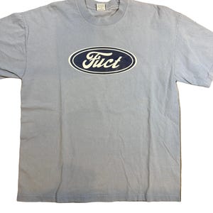 May include: A light blue t-shirt featuring a white and navy blue oval logo with the word "Fuct" in a stylized font. The shirt has short sleeves and a crew neck. The fabric appears to be a soft cotton blend.