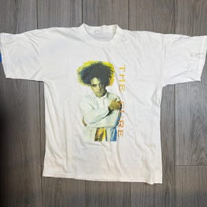 Op de afbeelding: Wit T-shirt met een portret van een man met donker, krullend haar. De woorden "THE CURE" staan verticaal aan de rechterkant. De afbeelding staat in het midden van het shirt. Het shirt is gemaakt van een zacht materiaal.