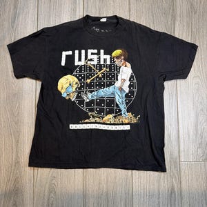 May include: Black t-shirt featuring the band name "RUSH" in white block letters. The graphic depicts a figure with a skull head, playing drums, surrounded by dice and bones. The text "ROLL THE BONES" is below the graphic.